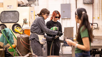 One person is holding a tool and talking to another over a workdesk.