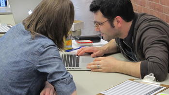 Two people lean across the table to look at something on a computer.