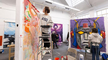 A student in a RISD shirt is on a ladder painting a large piece of art.