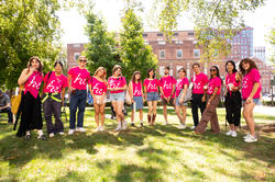 A group of RISD orientation leaders in pink shirts with a print that says "Hi" standing on a green with trees and a brick campus building behind them.